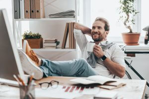 Handsome young man holding one hand behind head and holding a cup with another hand while sitting on working place in creative office