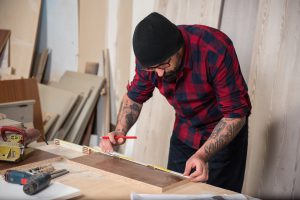 Adult man with beard and tattoo working in carpenter workshop using power tools