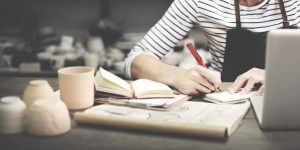 Arm of a woman sitting at a desk with sleeve pushed up, pencil in hand, notebook open and plans laid out on the desk