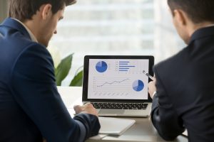 Rear view at two businessmen analyzing stats financial data on pc laptop, pointing at screen with rising graph and charts, discussing company growth, using easy accounting software for small business