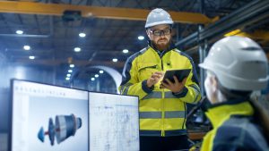 Inside the Heavy Industry Factory Female Industrial Engineer Works on Personal Computer She Designs 3D Engine Model, Her Male Colleague Talks with Her and Uses Tablet Computer.